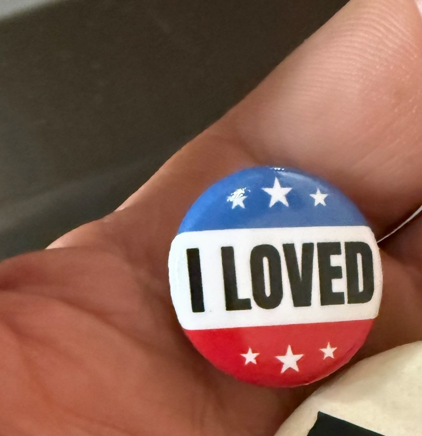 A red, white, and blue campaign-style pin that reads I LOVED, held in a hand