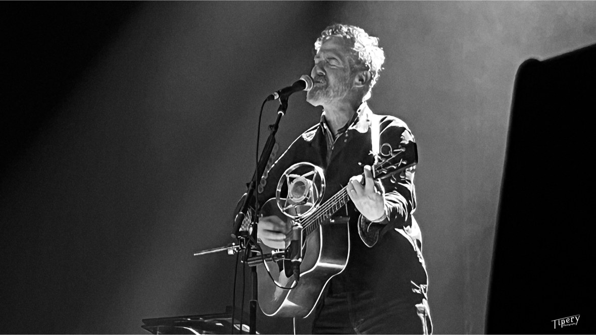 Glen Hansard playing acoustic guitar and singing into a microphone on stage, in black and white